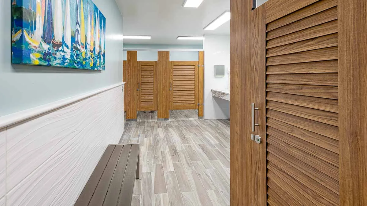 Marina bathroom features compact laminate, wood grain louver doors in headrail braced style. Sailboats print on wall above grey metal changing bench.