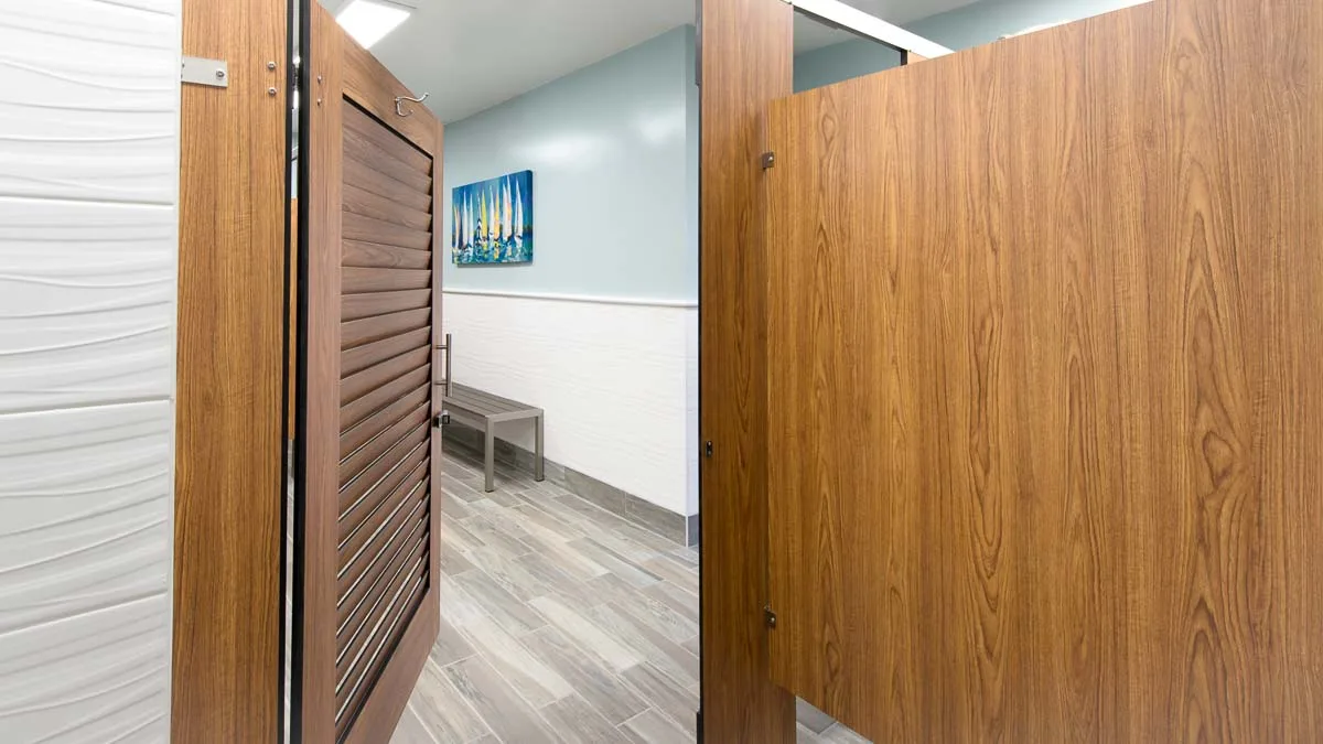 Inside compact laminate, wood grain dressing compartment looking out open louver door into marina bathroom. Sailboat print and metal changing bench.