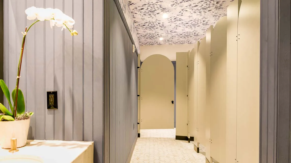 Unique hotel bathroom with custom shape, rounded tombstone top of doors. Cream colored laminate stalls in grey bead board room with orchid on vanity.