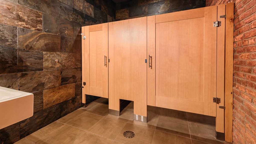 Earthy, urban bathroom features honey colored wood veneer partitions with two captured panel doors in floor mount style. Brick and slate walls.