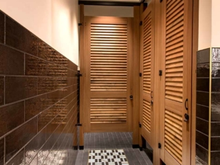 Trendy hotel bathroom with high privacy laminate wood grain partitions and three louver doors with powder coat hardware in dark subway tile bathroom.