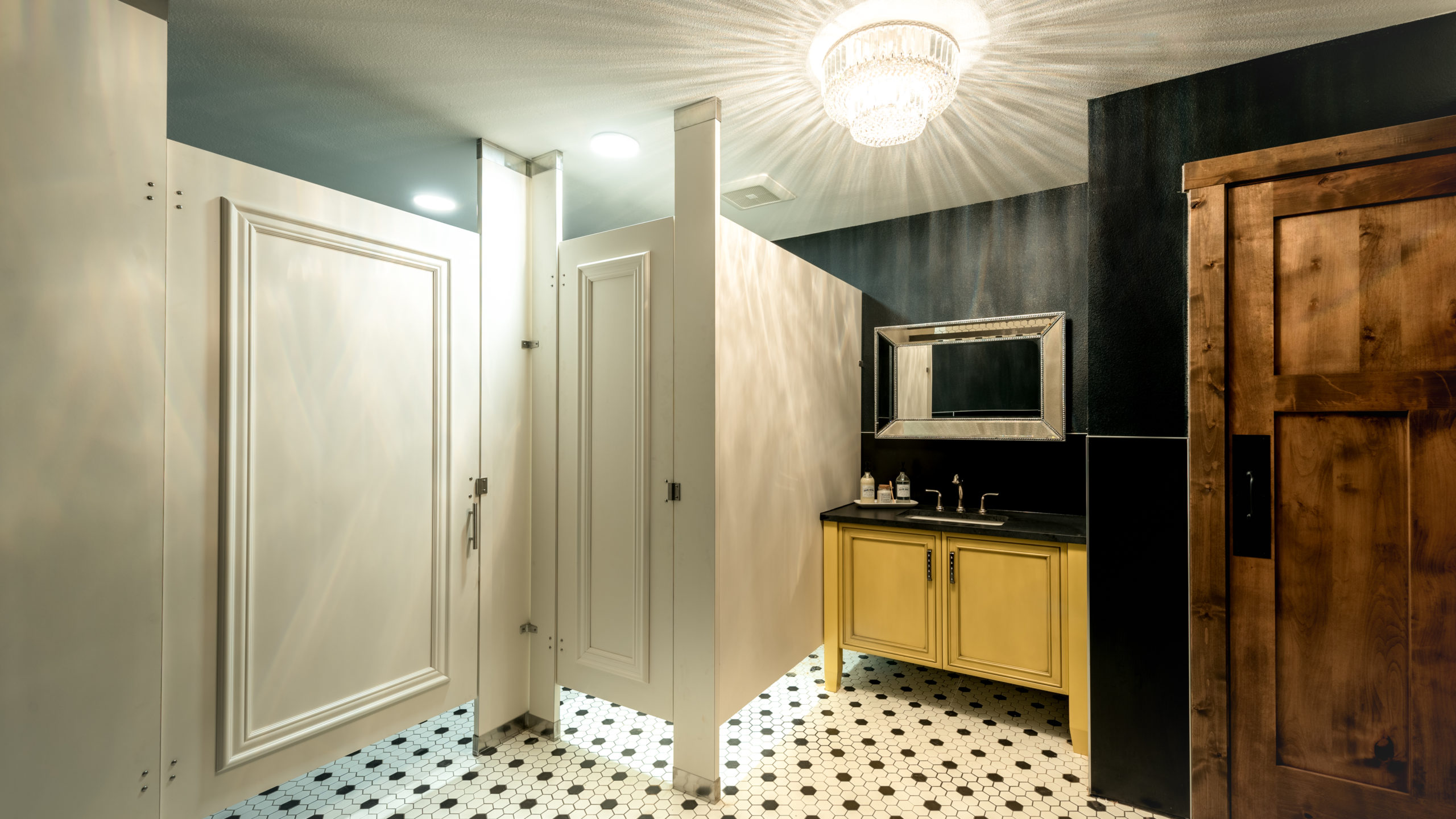 Designer showroom bathroom with white laminate partitions show doors with picture frame molding. Yellow vanity, silver framed mirror and chandelier.