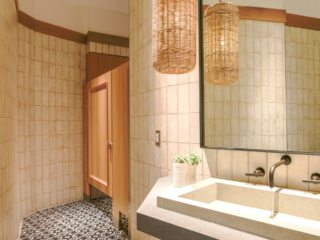 Trendy restaurant bathroom features tan tiled walls with one wood veneer partition and picture frame molding on door. Mirror and modern quartz sink.