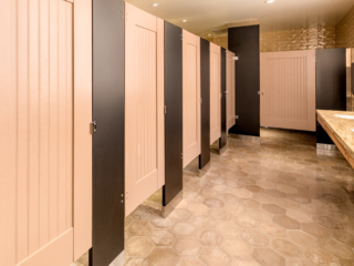 Large hotel bathroom featuring laminate partitions and six tan painted captured panel doors with bead board insert and dark brown pilasters.