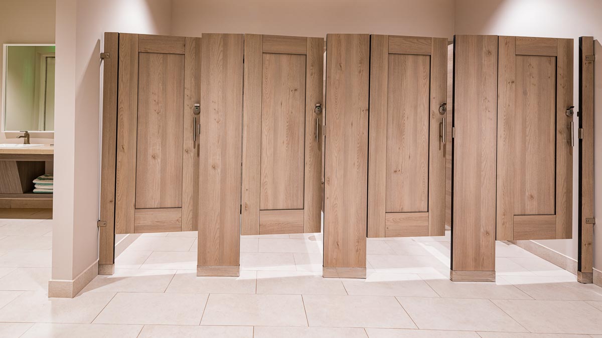 Country Club bathroom featuring compact laminate partitions with light tan, wood grain captured panel on slightly opened doors. Vanity on left.