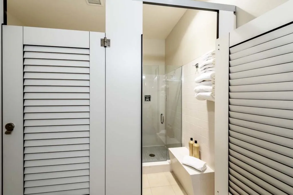 Two pretty, white compact laminate dressing compartments with louver doors. Open door shows changing area with bench leading to clear shower door.