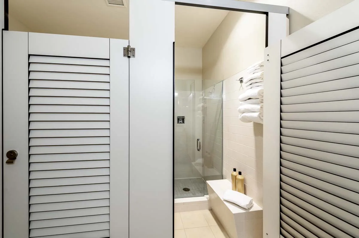 Two pretty, white compact laminate dressing compartments with louver doors. Open door shows changing area with bench leading to clear shower door.