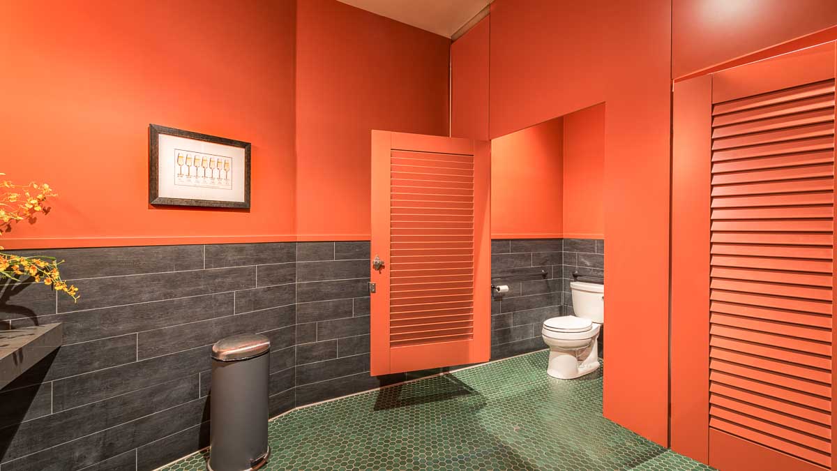 Winery bathroom painted bright orange in ceiling hung style with two louver doors and transom. Octagonal floor tile and wine glass print on wall.