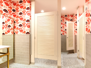 Trendy restaurant galleria bathroom shows wallpaper full of large red lips, cream subway tiles on wall showcases bright white laminate louver doors.
