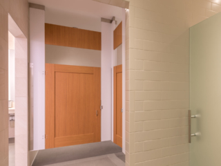 White subway tiled trendy business gym bathroom shows ceiling hung honey colored, wood grain captured panel doors with transom. Frosted shower door.
