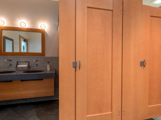 Craftsman style bathroom shows honey colored wood veneer partitions with two captured panel doors next to stone vanity and wood framed mirror.