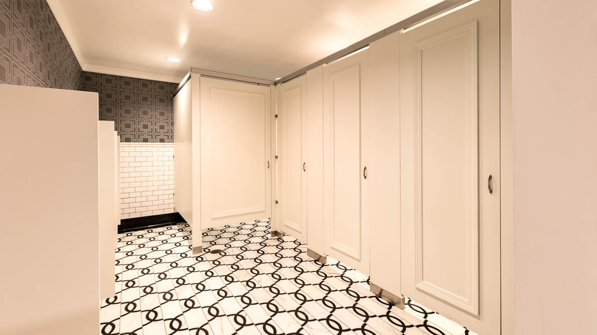 Grand, bright white hotel men’s bathroom with four doors showing picture frame molding in headrail braced style. Geometric design on walls and floors.