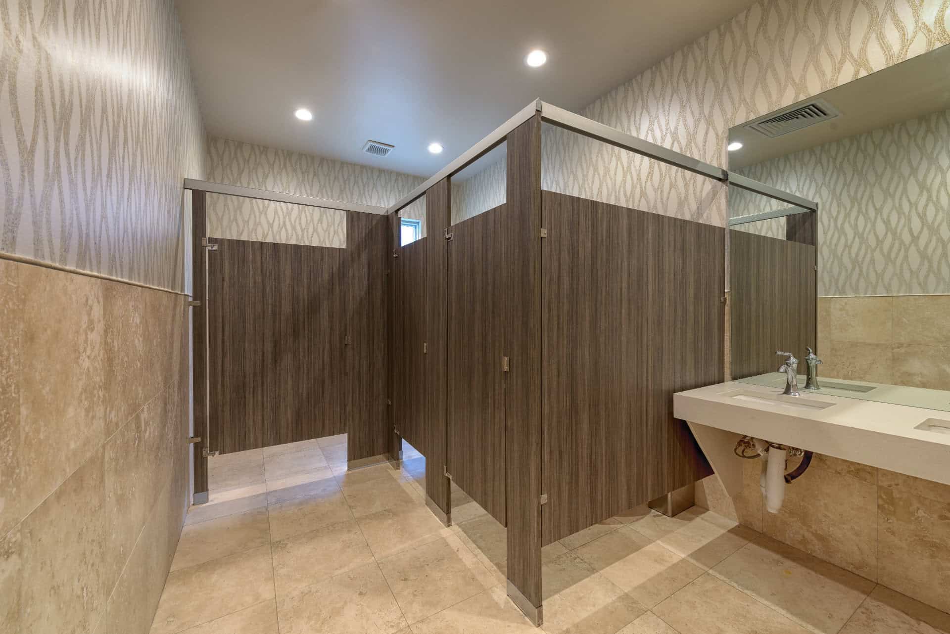 A modern headrail braced restroom with tiled flooring and walls, featuring three enclosed stalls and a handwashing sink with a single faucet.