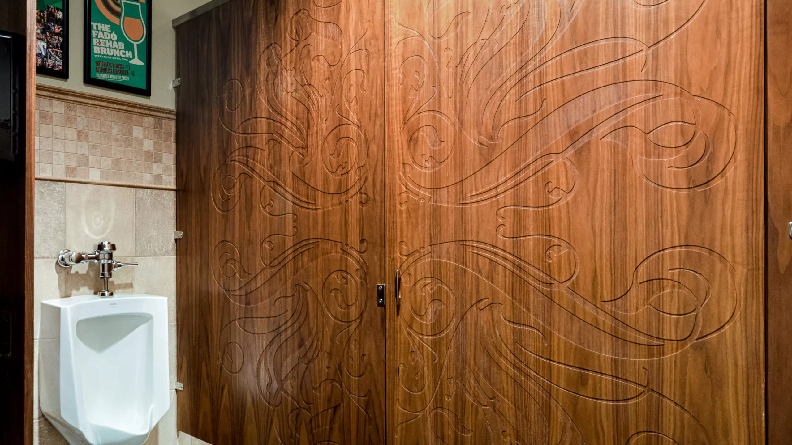 A picture of a public restroom wood veneer toilet partition with intricately engraved door and panel. There is a urinal on the left and two framed posters on the wall.