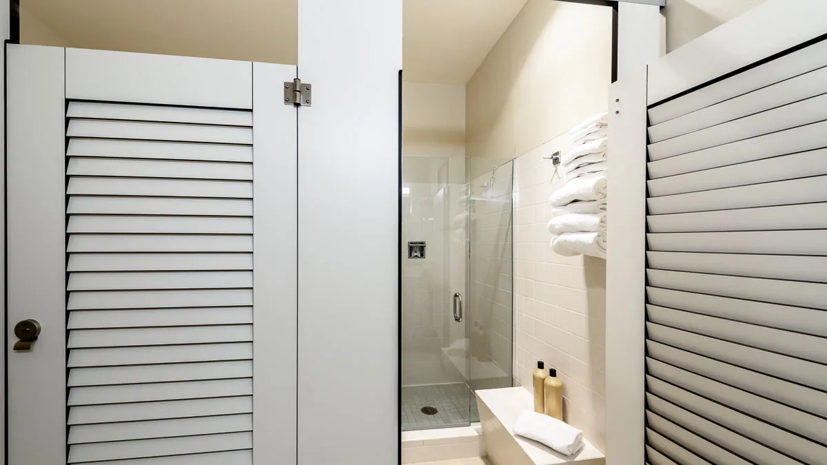 Two pretty, white compact laminate dressing compartments with louver doors. Open door shows changing area with products on bench and rolled towels.