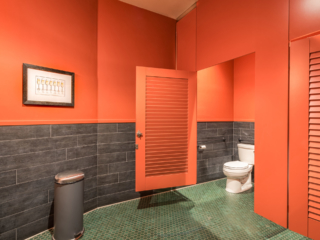 Winery bathroom painted bright orange in ceiling hung style with two louver doors and transom. Octagonal floor tile and wine glass print on wall.