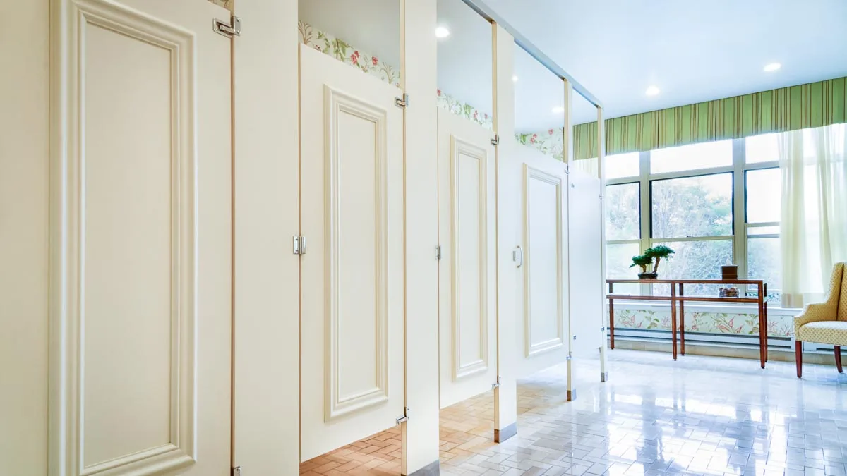 Beautiful white public restroom partitions with rectangular moldings on the doors and large, sunlit windows at the back of the picture. The partitions are made with high pressure laminate and are headrail braced.