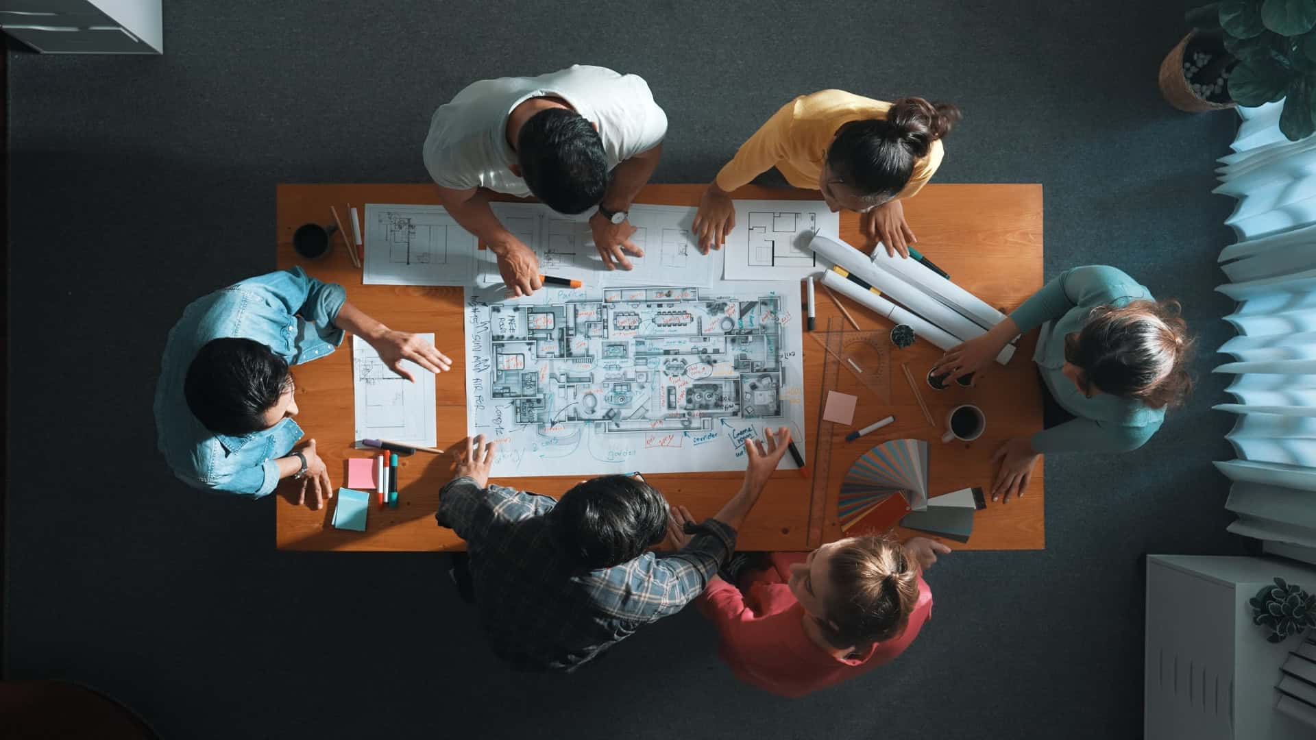 A diverse group of people work collaboratively around a table covered in architectural plans, color samples, and stationery, in a well-lit office space.