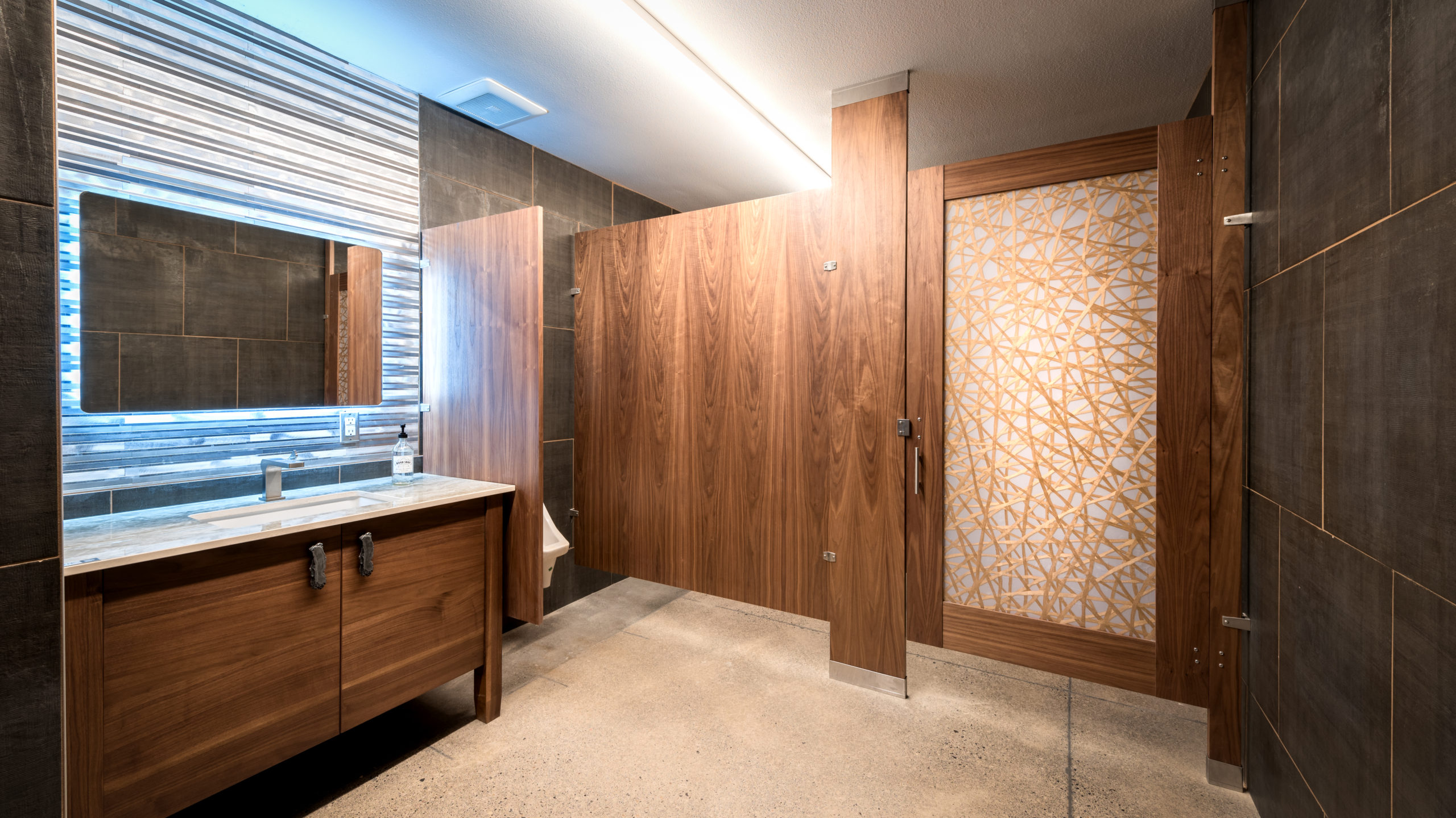 Dark rich wood veneer bathroom partition and acrylic insert door showing stray crossing gold lines and corrugated metal backsplash and cabinet vanity.