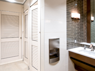 Dealership showroom bathroom with high privacy European style toilet partition shows white laminate louver doors and transoms. Brown tile urban room.