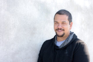 A man with short hair and a beard, wearing a dark hoodie, stands against a light-colored wall, smiling slightly.