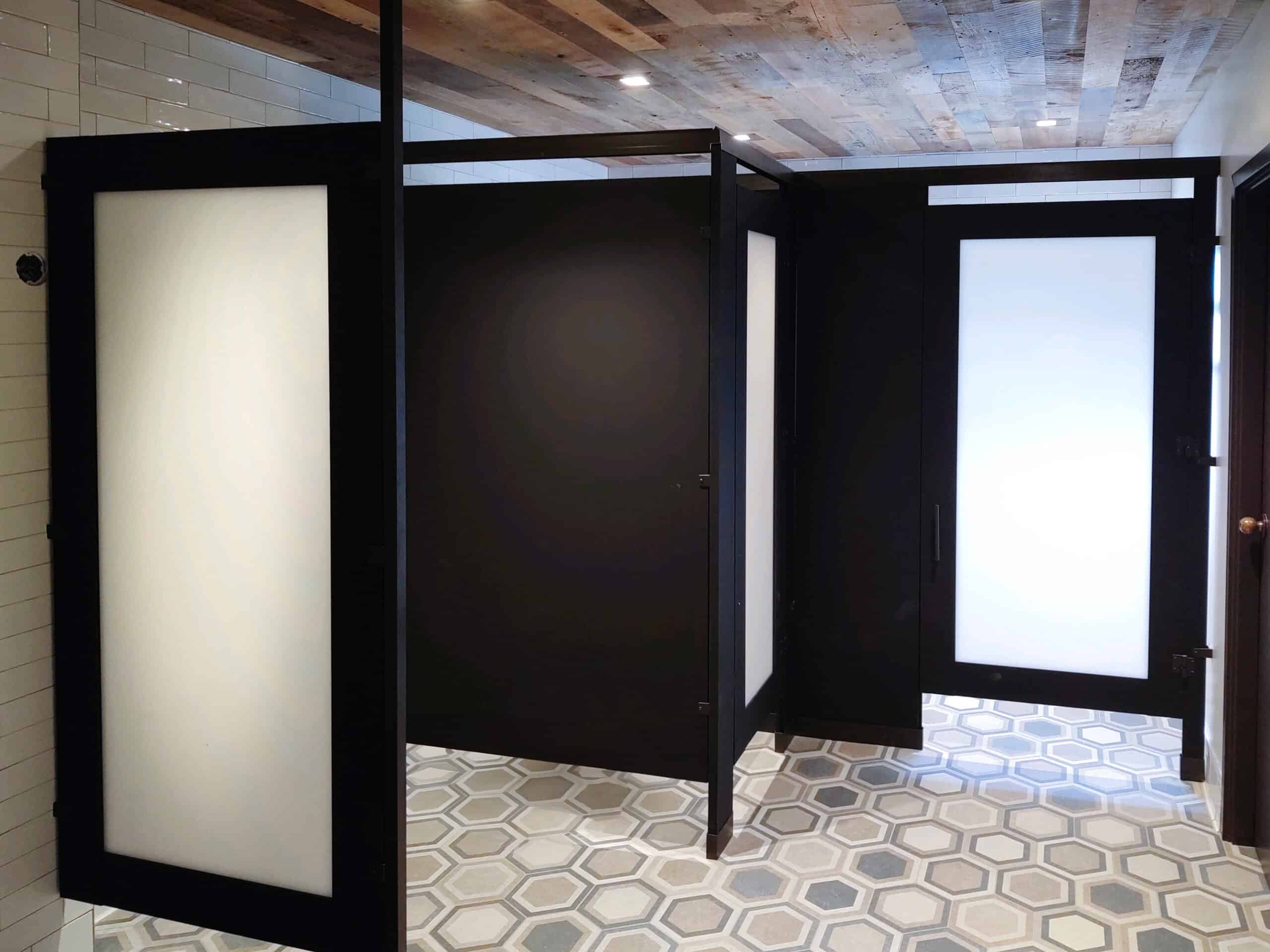 A beautiful public restroom featuring color black stall partitions, frosted glass doors, hexagonal tile flooring, and a wood-paneled ceiling.