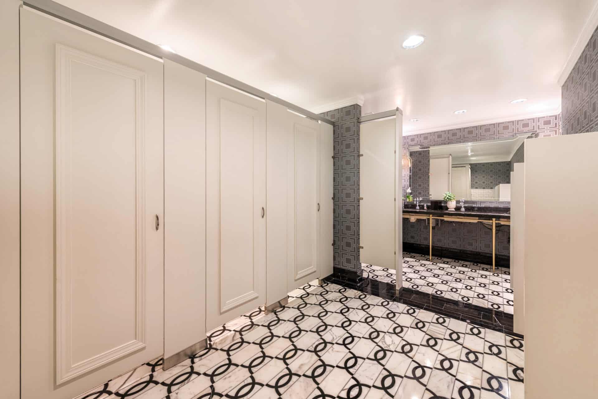 A public restroom with cream-colored Zero Sightline stalls, patterned tile floor, decorative wallpaper, and a counter with a large mirror and sinks offers a modern privacy solution for commercial restroom partitions.