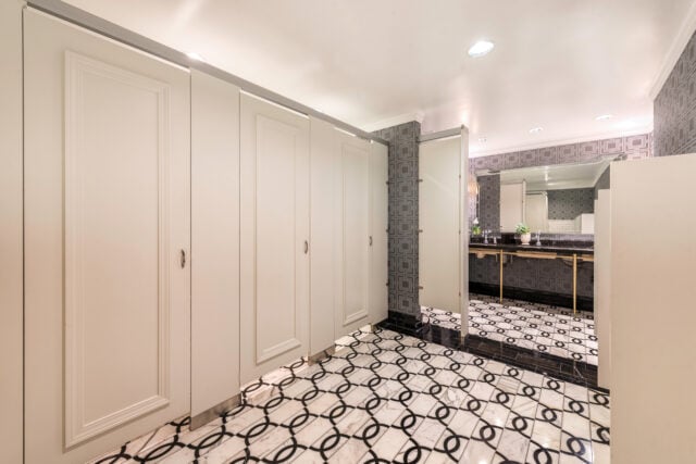 A clean public restroom with white stalls, patterned wallpaper, and black-and-white tile flooring. A mirror reflects a vanity with a sink and flowers.