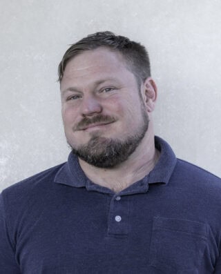 A man with short hair and a beard is wearing a dark blue polo shirt and standing in front of a plain light-colored background.