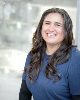 A person with long, wavy hair smiles while wearing a blue shirt. The background is softly blurred.