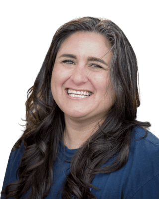A person with long, wavy dark hair smiles directly at the camera, wearing a dark top, against a plain background.