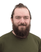 A person with a beard and long hair tied back is wearing a green shirt, standing against a plain background.