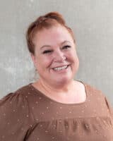 A smiling woman with reddish-brown hair tied up, resembling Janice Gaynor, wears a brown blouse with small patterns as she stands against a textured gray background.
