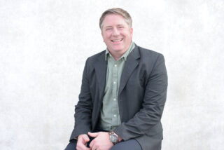 A man in a gray blazer and green shirt is sitting and smiling against a light-colored, textured background.