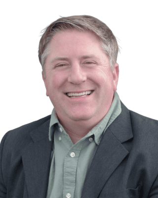 A person with short light hair is smiling, wearing a green collared shirt and dark blazer, against a plain background.