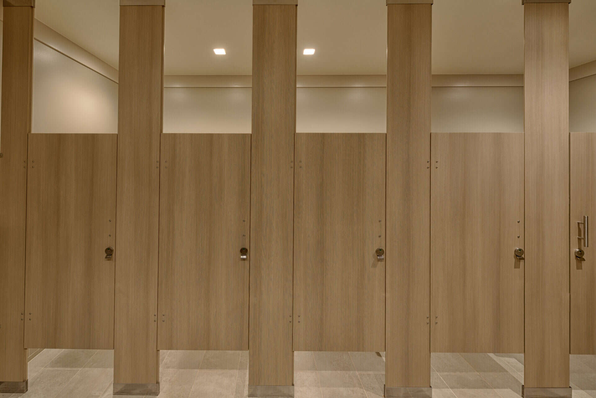 Row of closed wooden restroom stall doors in a public bathroom, featuring commercial restroom partitions as a privacy solution, with tiled floor and overhead lighting.