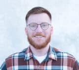 Eljay Johnson, a man with short red hair, a beard, and round glasses, is smiling at the camera. He is wearing a plaid shirt and standing in front of a light-colored background.