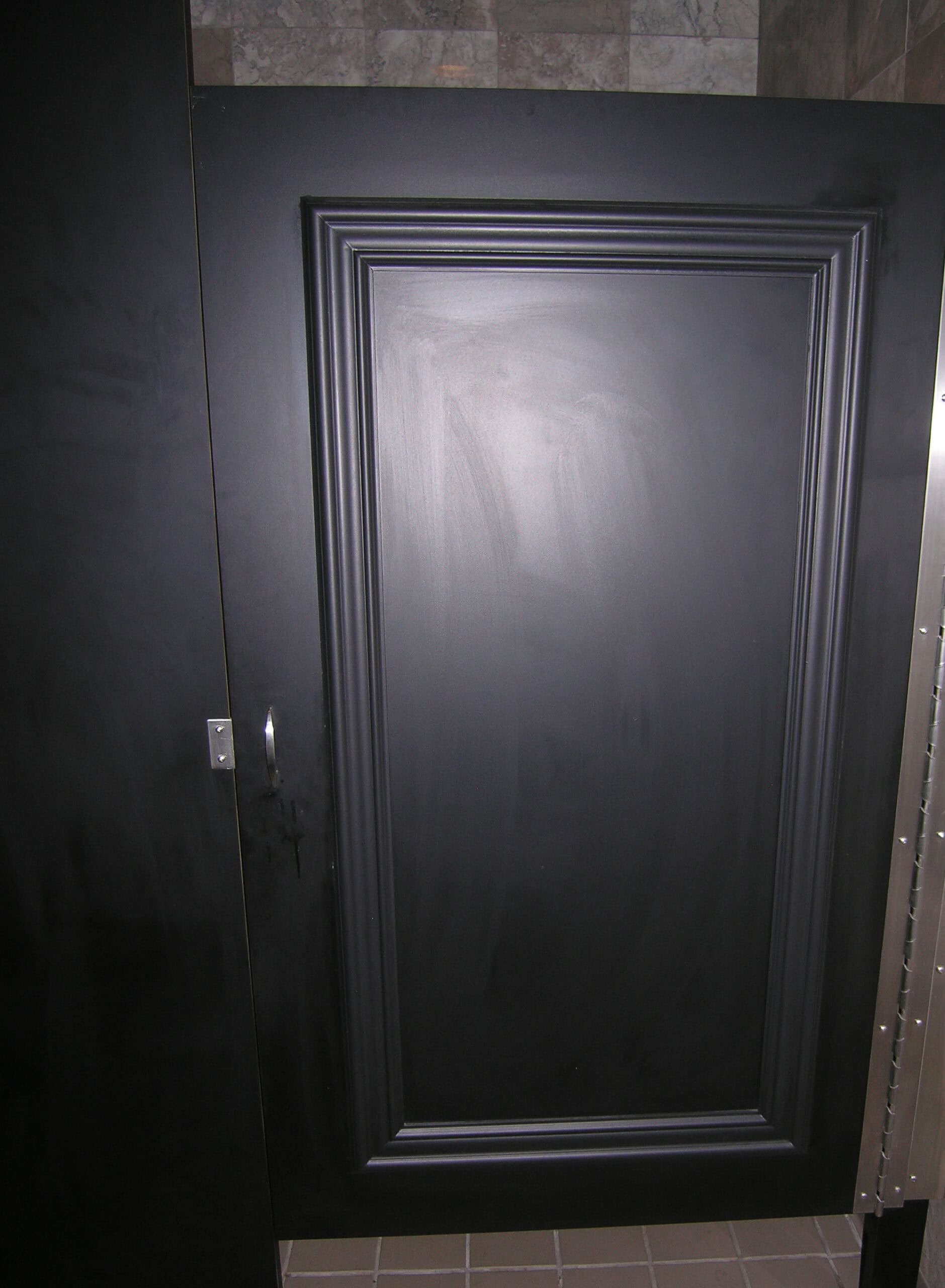 A beautiful closed bathroom stall door in color black features a rectangular recessed panel, metallic handle, and tiled floor visible below.