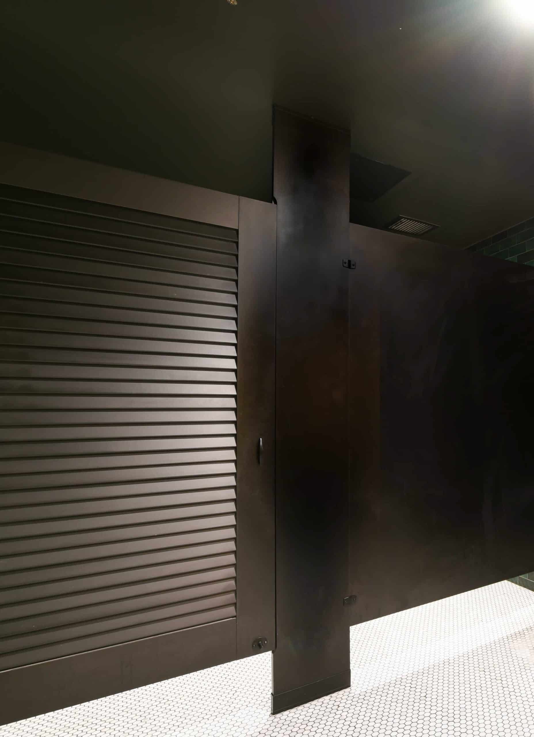 A dark restroom stall featuring the beautiful color black, with a closed metal shutter on one side and a solid black partition, set on a white tiled floor.