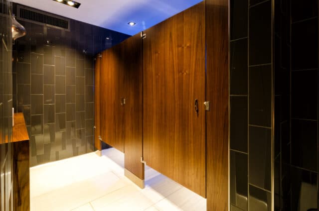 A public restroom with two wooden stall doors, black tiled walls, and overhead lighting.