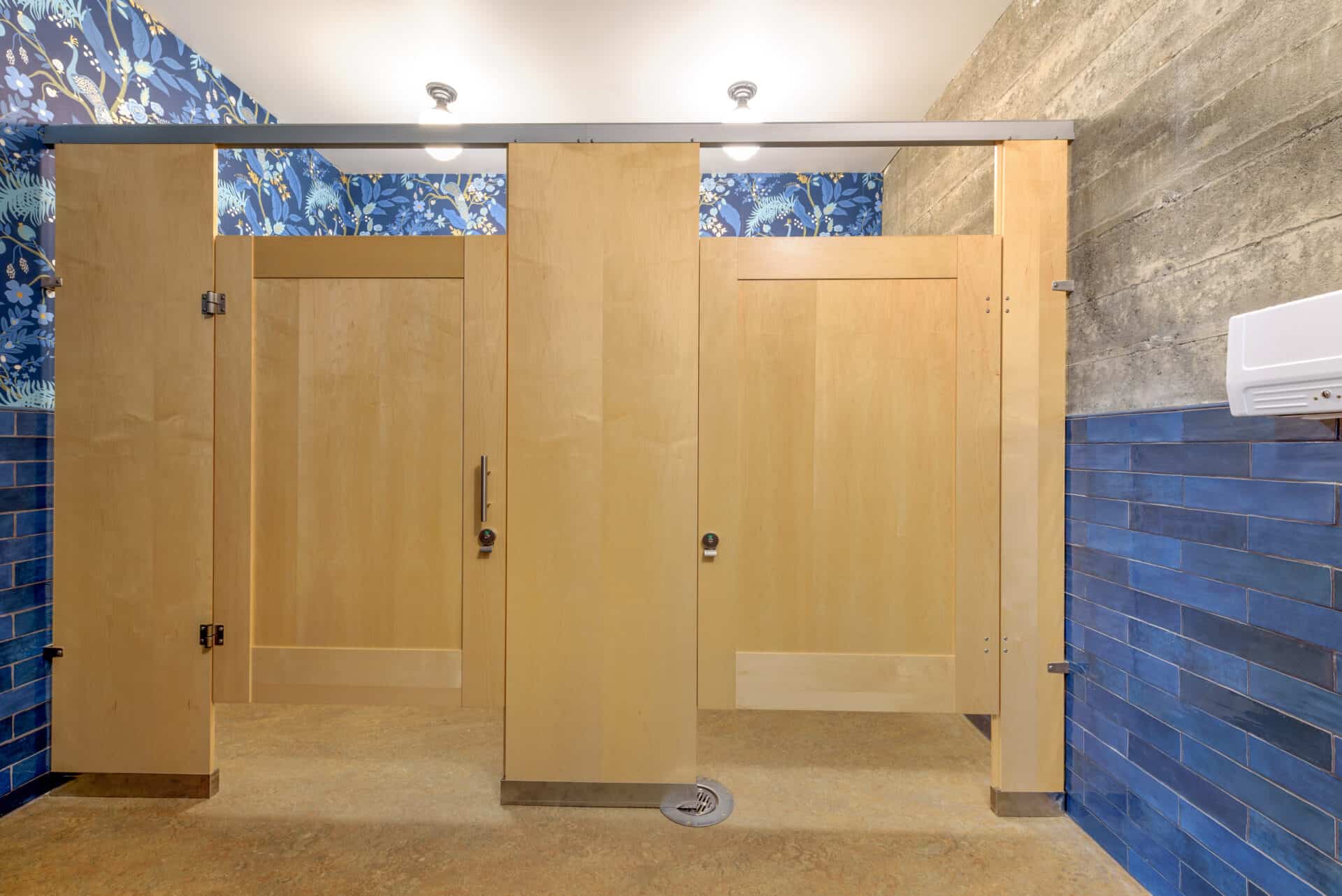 Three wooden restroom stalls feature blue tile walls and floral wallpaper above, with a hand dryer on the right wall—an inviting space that showcases Commercial Restroom Partitions for a stylish privacy solution.