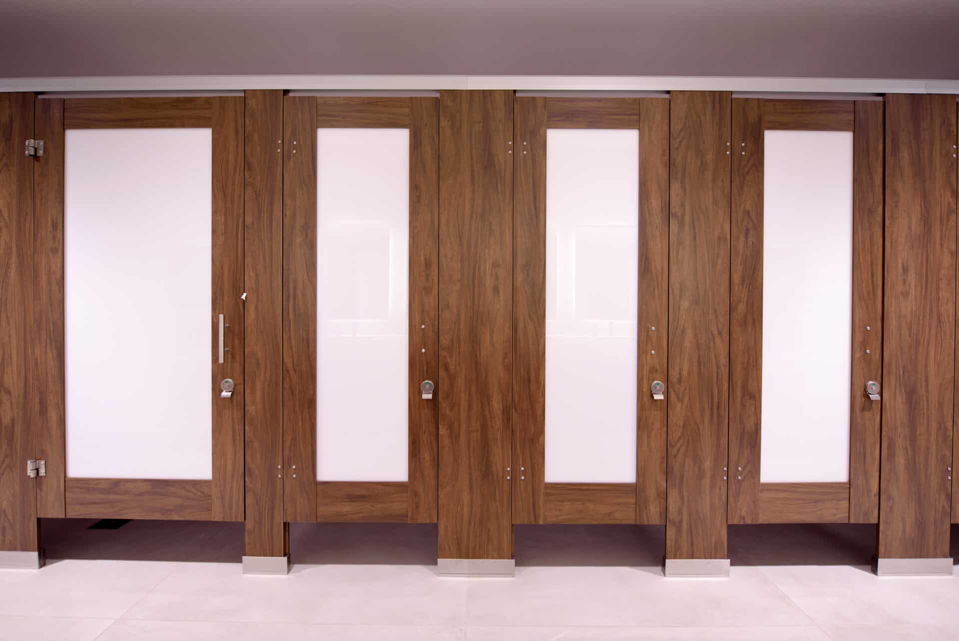 Row of closed wooden restroom partitions with frosted glass panels and silver locks on a tiled floor, featuring a zero sightline privacy solution.