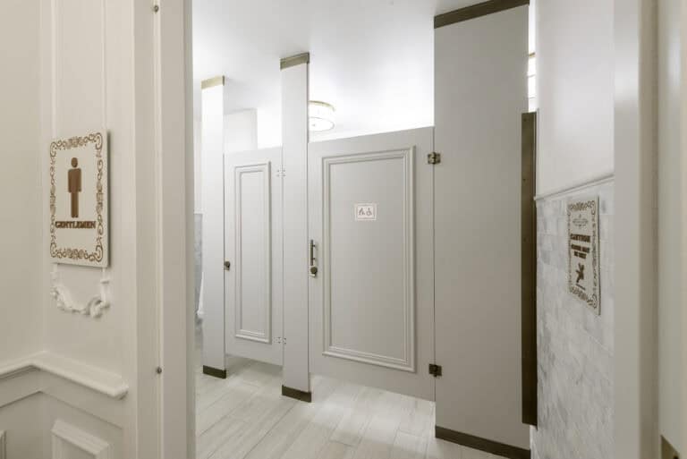 Elegant restroom partitions featuring classic paneled doors in a cream finish with bronze hardware and trim, extending to the ceiling with upper transoms for added privacy, under soft overhead lighting.