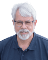 An older adult with short white hair and glasses, wearing a dark polo shirt, is smiling.