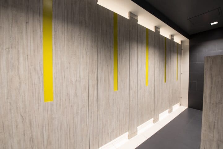 Restroom partitions made from high-pressure laminate with a light woodgrain pattern, bold yellow acrylic inlays, and a floor to ceiling mountings, creating a modern and vibrant design for commercial restroom.