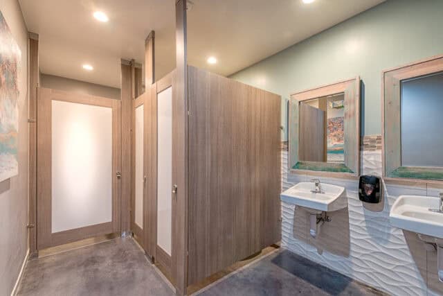 Commercial restroom partitions made from woodgrain plastic laminate, featuring floor to ceiling mounting, high privacy, and frosted acrylic door lites for a modern and functional design.