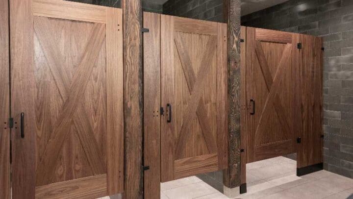Three closed wooden restroom stalls with dark handles, set against tiled flooring and gray brick walls.