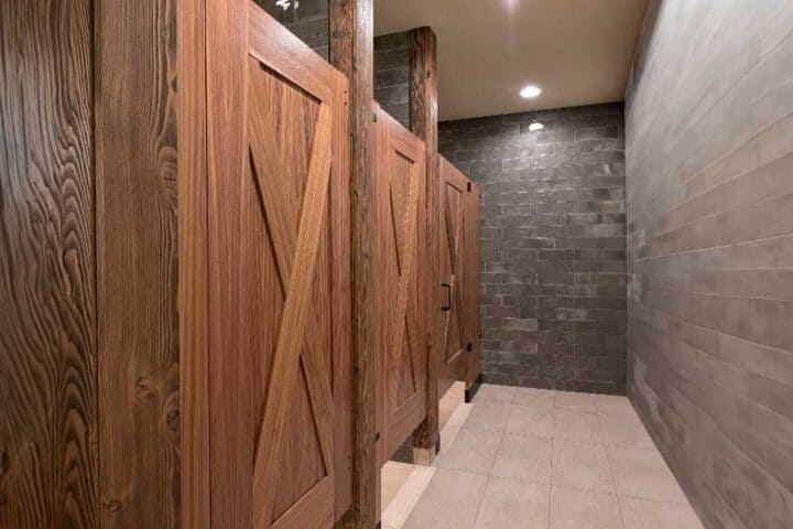 A public restroom with wooden stall doors and tiled walls and floor, illuminated by ceiling lights.