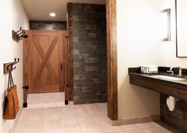 Modern restroom with a wooden stall door, dark tile walls, a countertop sink with a mirror, and hooks holding a bag on the left wall.