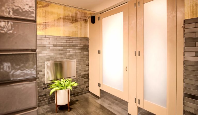 A photo of restroom partitions with frosted acrylic door lights mounted floor to ceiling with a plant at the lower left of the image.
